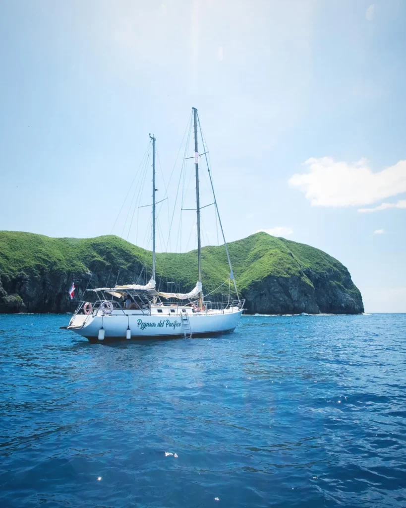 Pegasus sailboat with sails lowered beside a green rocky headland in Guanacaste